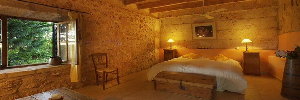 A bedroom at Sant Blai: bare mares stone walls, a white bed between two lamps, a deep stone window onto the garden