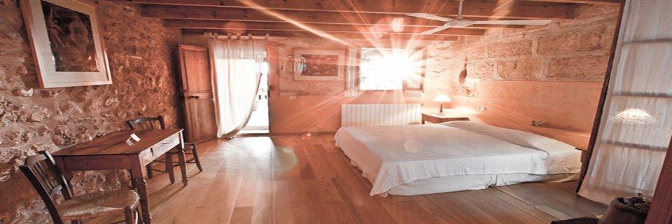 A bedroom at Sant Blai with mares walls, a wood floor and a white bed, afternoon light flaring through the open doorway